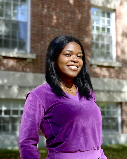 A girl with dark hair is smiling at the camera, wearing a vibrant purple sweater and posed in front of a brick background.