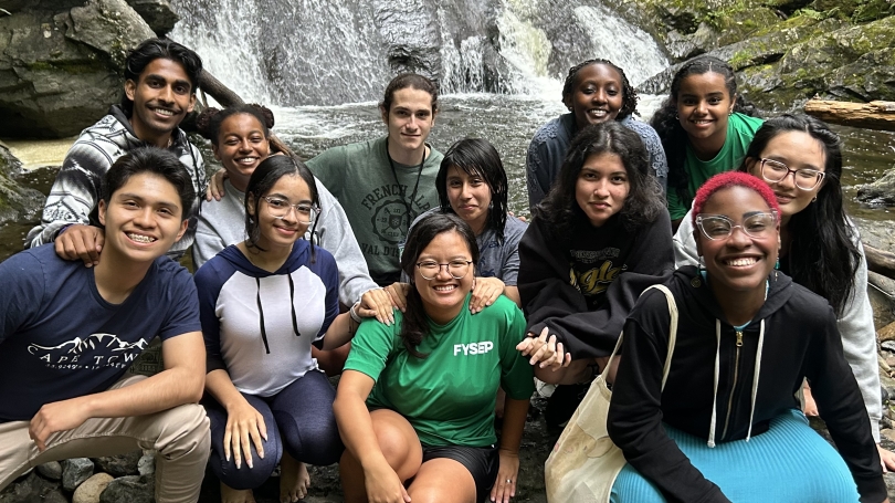  A group of smiling students posing together on a large rock in front of a picturesque waterfall, surrounded by lush green trees.