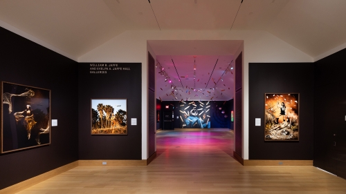 A museum gallery with dark brown walls and large scale framed photographs of Native American peoples leads into another gallery with dark gray walls, colored lighting, and corn suspended from the ceiling.