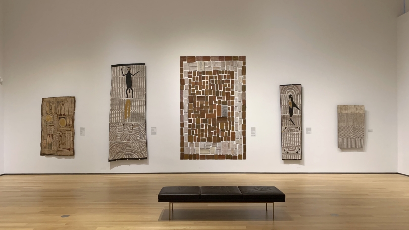 A museum gallery with a high ceiling and white walls. On the wall are four large works of Indigenous Australian bark paintings.