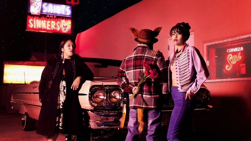 Cara Romero's "Coyote Tales No. 1" Three figures stand confidently near a vehicle. One is facing away from the cara and holding flowers behind their back.
