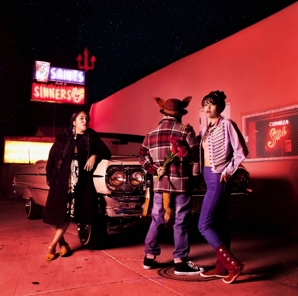Cara Romero's "Coyote Tales No. 1" Three figures stand confidently near a vehicle. One is facing away from the cara and holding flowers behind their back.