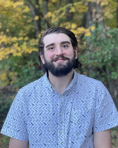 Dark haired, fair skinned male with a dark beard and a smile, wearing a light blue patterned shirt, standing in front of fall foliage
