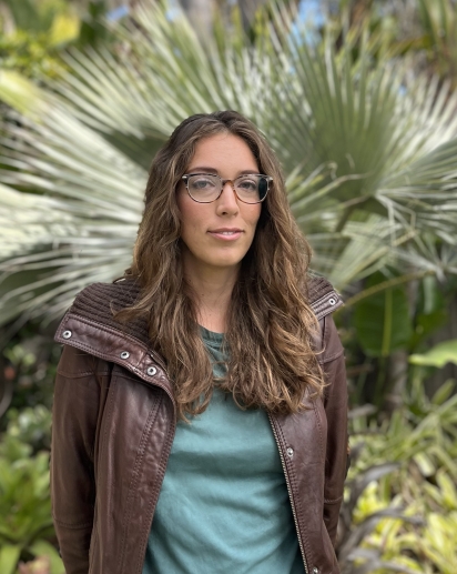 A tan woman with long light brown wavy hair, wearing glasses and a leather jacket, standing in front of palm trees