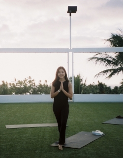 Rachel in black outfit in namaste pose, with yoga mat behind and greenery and palm in background
